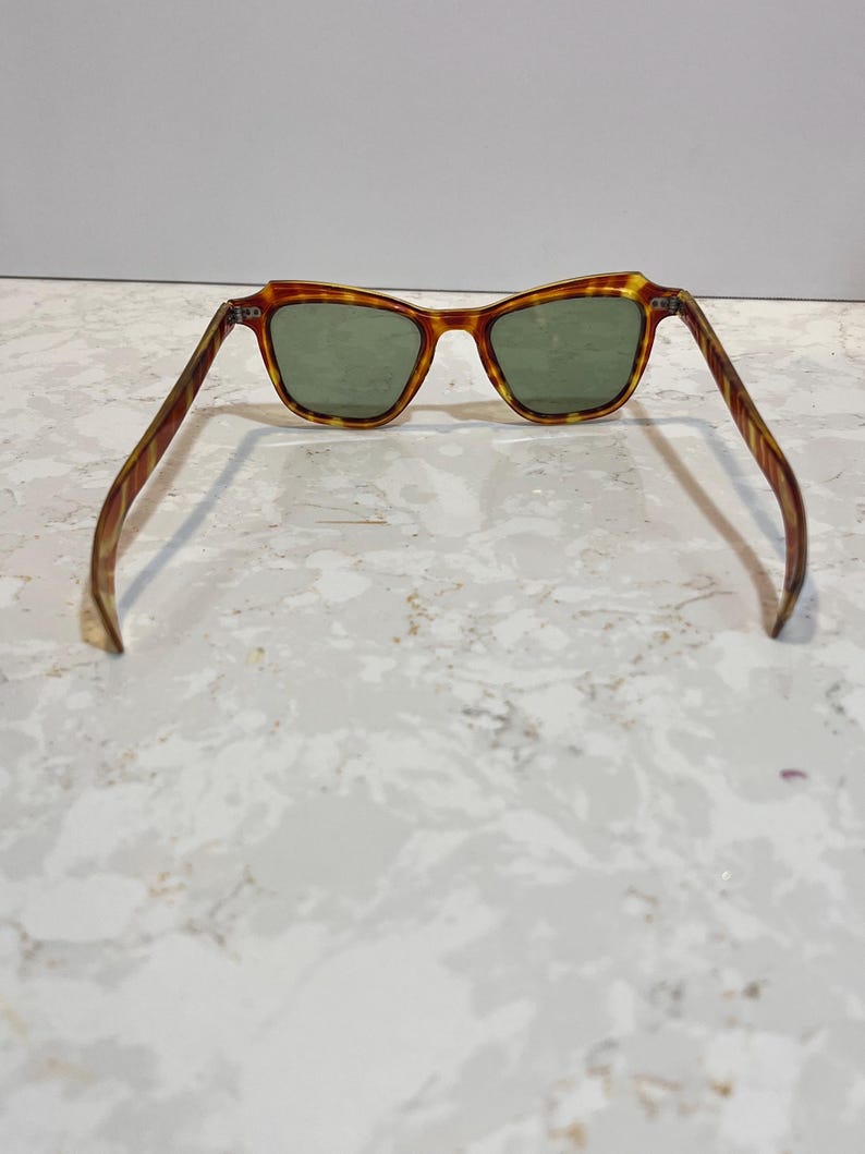 May include: Sunglasses with a tortoiseshell frame and green lenses. The frame has a brown and yellow pattern. The sunglasses are open, with the arms extended. The lenses are a dark green color.