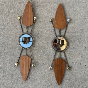 May include: Pair of mid-century modern wall sconces. Each sconce features a wooden shield shape, a circular mirrored element, and brass and silver metal accents. The sconces have a symmetrical design with a retro aesthetic.