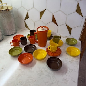 Könnte beinhalten: Ein Vintage-Teeservice aus orangefarbenem Kunststoff mit passendem Krug, Tassen, Untertassen und Schalen. Das Set umfasst verschiedene Farben wie Orange, Gelb, Grün und Braun. Die Artikel sind auf einer weißen Arbeitsplatte angeordnet.