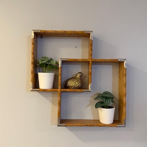 Puede incluir: Una estantería de pared de madera que consta de dos marcos cuadrados interconectados con bordes festoneados y esquinas de metal plateado. Dos macetas blancas pequeñas con plantas verdes y una figura de pájaro dorada están dentro.