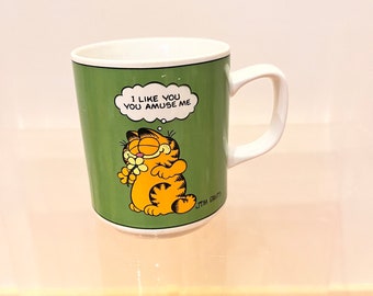 May include: Green ceramic mug with a cartoon image of Garfield the cat holding a flower. The mug has the text "I like you you amuse me" in a thought bubble above Garfield's head.