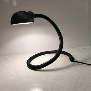 Peut inclure: Une lampe de bureau noire avec un col flexible et enroulé et un abat-jour arrondi. La lampe est allumée, projetant une lumière vive. La base n'est pas visible. Le design de la lampe est moderne et minimaliste.