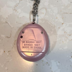 May include: A pink, handheld electronic game keychain. The back of the device has the text "BANDAI 1997 MADE IN CHINA". The keychain has a silver metal chain and ring.
