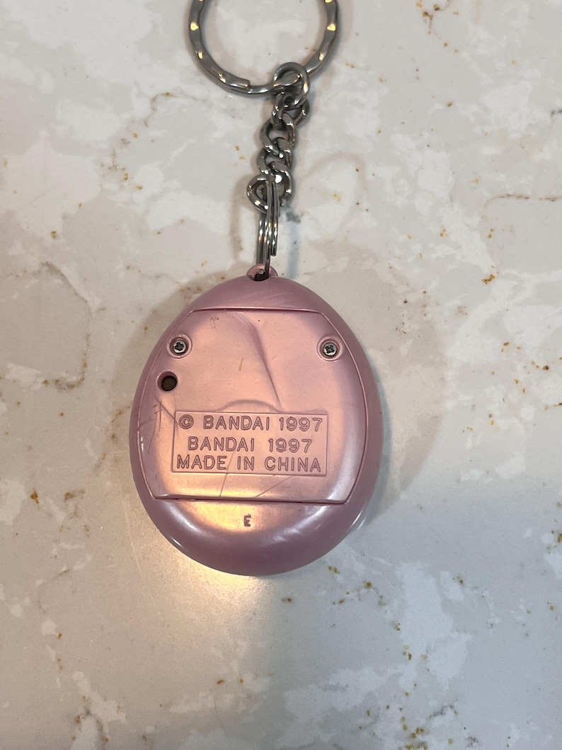 May include: A pink, handheld electronic game keychain. The back of the device has the text "BANDAI 1997 MADE IN CHINA". The keychain has a silver metal chain and ring.