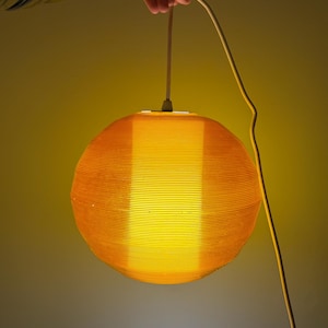 May include: A spherical orange pendant lamp with a ribbed texture, radiating a warm yellow light. The lamp is suspended by a white cable, held by a hand. The design is modern and minimalist, suitable for interior lighting.