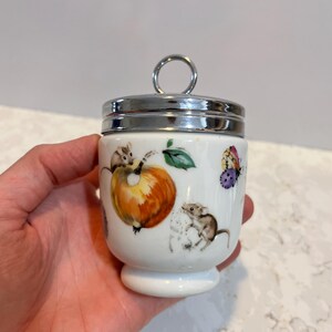 May include: White ceramic egg cup with a silver lid and a whimsical design featuring mice, fruit, and butterflies.