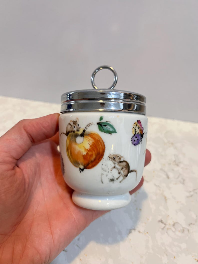 May include: White ceramic egg cup with a silver lid and a whimsical design featuring mice, fruit, and butterflies.