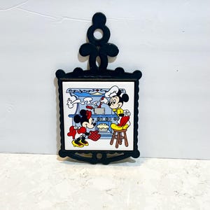 May include: A black cast iron pot holder with a white ceramic tile featuring Mickey and Minnie Mouse cooking in a kitchen. Minnie is holding a red oven mitt and Mickey is stirring a pot on the stove.