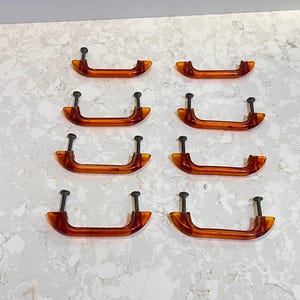 Könnte beinhalten: Acht bernsteinfarbene Schrankgriffe mit schwarzen Schrauben sind auf einer weiß-grau gesprenkelten Oberfläche angeordnet. Die Griffe haben eine gebogene, rechteckige Form und bestehen aus einem durchscheinenden Material. Die Hardware ist wahrscheinlich für die Möbelrestaurierung oder ein DIY-Projekt gedacht.