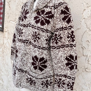 May include: A brown and white knit cardigan with a brown snowflake pattern. The cardigan has a shawl collar and a zipper closure.