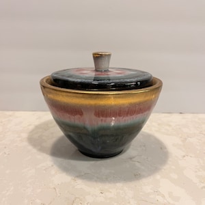 May include: A small ceramic jar with a lid. The jar is decorated with a multi-coloured glaze, including shades of pink, green, and black. The lid has a gold rim and a white knob.