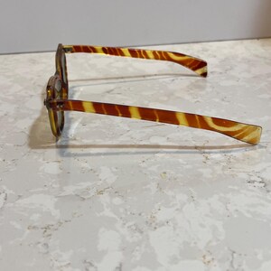 May include: A pair of vintage eyeglasses with a tortoiseshell pattern. The frames and arms are a mix of amber and brown, creating a striped effect. The glasses are resting on a light-colored surface.