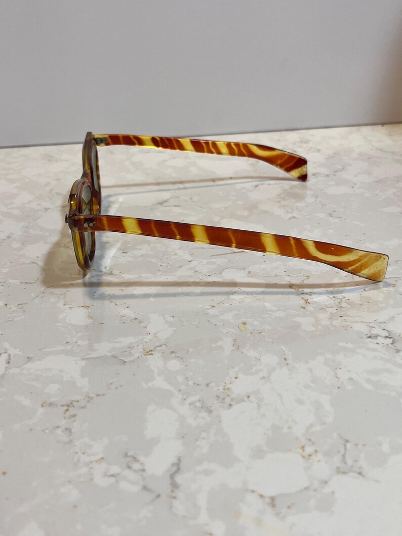 May include: A pair of vintage eyeglasses with a tortoiseshell pattern. The frames and arms are a mix of amber and brown, creating a striped effect. The glasses are resting on a light-colored surface.