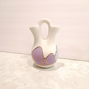 Peut inclure: Un vase double en céramique blanche avec une anse, décoré de motifs géométriques violets et dorés.
