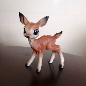 Könnte beinhalten: Keramikfigur eines Rehs mit großen, ausdrucksstarken Augen und einem süßen Ausdruck. Das Reh hat einen braunen Körper mit weißen Flecken und weißen Beinen. Die Ohren und die Spitzen der Hufe sind schwarz. Ein charmantes Dekorationsstück.