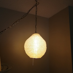 Op de afbeelding: Een witte, getextureerde, ronde hanglamp hangt aan een ketting. Het licht is aan en geeft een warm gloed.