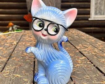 May include: A blue ceramic cat figurine wearing black framed glasses. The cat is sitting on a wooden surface with pine needles scattered around it.