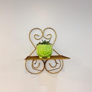 Può includere: Mensola da parete decorativa con design a forma di cuore in metallo color oro. La mensola contiene un piccolo vaso di ceramica verde con superficie strutturata e una pianta. La mensola è larga circa 25 cm.