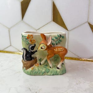 May include: A ceramic planter depicting a skunk and a fawn in a forest setting. The planter is rectangular with a light green background, orange flowers, and green leaves. The fawn is brown with white spots and the skunk is black and white.