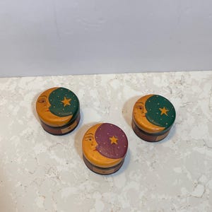 Könnte beinhalten: Drei kleine, runde Holzkisten mit Deckeln. Jede Box hat ein Mondgesicht-Design in Gelb- und Orangetönen. Die Deckel sind grün, lila und gelb mit einem Stern. Die Boxen haben einen dunkelbraunen Sockel.