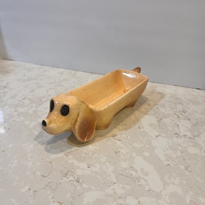 May include: A ceramic dachshund shaped dish with a light brown glaze. The dish has a small tail and black eyes.
