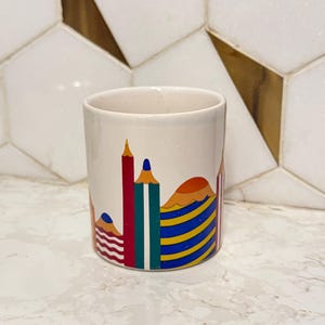 Può includere: Tazza cilindrica in ceramica con base color crema. La tazza presenta un design colorato di matite stilizzate e forme a strati in rosso, blu, verde, giallo e arancione. Adatta per bevande.