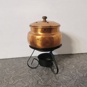 May include: A copper pot with a lid and a black metal stand. The pot has a small knob on the lid and several holes in the top. The stand has three legs and a small black dish in the centre.