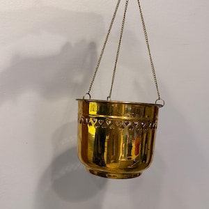 Peut inclure: Un pot de fleurs suspendu doré brillant avec un motif en forme de cœur sur le dessus. Le pot est suspendu par trois fines chaînes dorées. Le pot est tenu par une main sur un fond blanc.