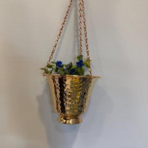 May include: A gold-coloured metal hanging planter with a hammered texture and a flared rim. The planter is suspended by three thin gold-coloured metal chains that meet at the top. Inside the planter are small green leaves and blue flowers.