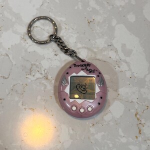 May include: A pink Tamagotchi Angel virtual pet with a silver keychain. The device has a small screen displaying a pixelated character. The text "Tamagotchi Angel" is above the screen, and there are three buttons below.