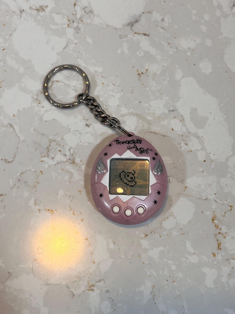 May include: A pink Tamagotchi Angel virtual pet with a silver keychain. The device has a small screen displaying a pixelated character. The text "Tamagotchi Angel" is above the screen, and there are three buttons below.