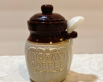 Könnte beinhalten: Eine Keramikdose mit braunem Deckel und einer weißen Schaufel, die an der Seite befestigt ist. Die Dose ist mit einem braunen und weißen Design und den Worten "INSTANT COFFEE" in einer dekorativen Schrift verziert.