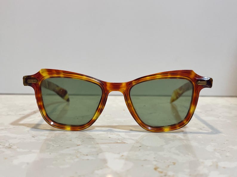 May include: Vintage-style sunglasses with a tortoiseshell pattern frame and green lenses. The frames have a classic, slightly angular shape. The sunglasses are designed for eye protection and fashion.