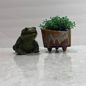 Könnte beinhalten: Eine grüne Keramik-Froschfigur sitzt neben einem braunen und weißen Keramiktopf mit einer kleinen grünen Pflanze darin. Der Topf hat drei Beine.