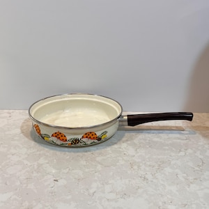 Può includere: Una padella vintage color crema con un manico marrone scuro. La padella presenta un bordo decorativo con illustrazioni di funghi arancioni e marroni. La padella è posta su uno sfondo neutro.