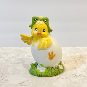 May include: A yellow ceramic chick with a green bow peeks out of a white egg shell. The chick is sitting on a bed of green grass with white flowers.
