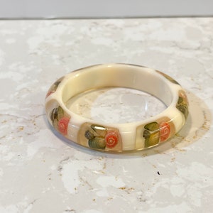 May include: White bangle bracelet with a design of seashells and flowers in shades of pink, green, and brown.
