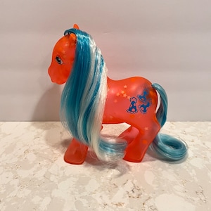 Könnte beinhalten: Ein leuchtend orangefarbenes My Little Pony mit blauem und weißem Haar. Das Pony hat ein Sternen- und Schmetterlingsdesign an der Seite.
