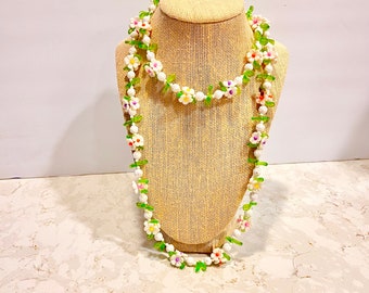 May include: A white and green beaded necklace with white flower-shaped beads and green leaf-shaped beads. The necklace is displayed on a beige fabric mannequin.