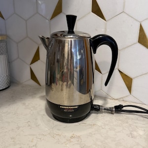 May include: A stainless steel Farberware percolator with a black base, handle, and lid knob. The electric coffee maker sits on a white countertop with a geometric tile backsplash. The Farberware logo is visible on the front.