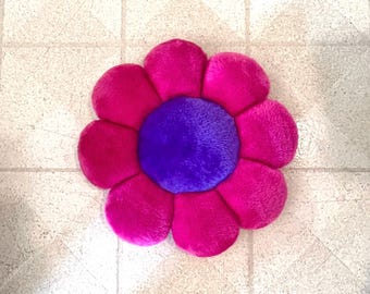 May include: A plush pink flower cushion with a purple center. The cushion is made of soft, fuzzy fabric and has a round shape.