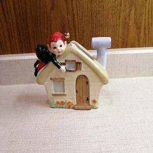May include: A ceramic figurine of a house with a green roof and a chimney. A small elf with red hair and a green outfit is climbing on the roof.