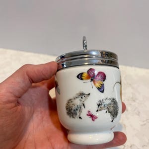 May include: White ceramic egg cup with a silver lid and handle. The cup features a colorful butterfly and two hedgehogs with a pink butterfly.