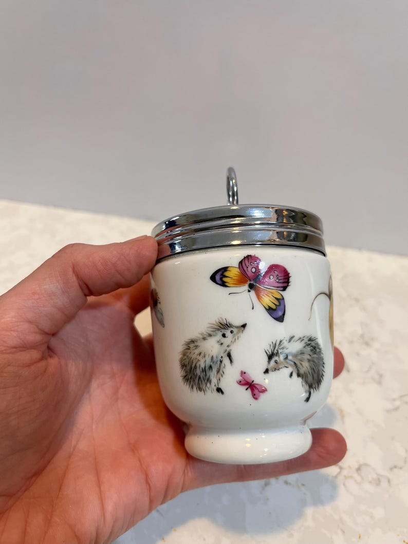 May include: White ceramic egg cup with a silver lid and handle. The cup features a colorful butterfly and two hedgehogs with a pink butterfly.