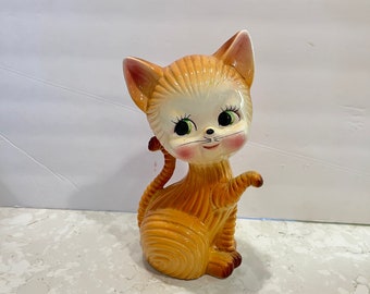 May include: A ceramic figurine of a cat with large, expressive eyes. The cat is sitting with its right paw raised, and its body is painted in shades of brown and yellow.