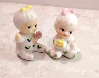 May include: Two small porcelain figurines of children dressed in pajamas. The figurine on the left is wearing a yellow bow and has a green and white floral pattern on their pajamas. The figurine on the right is wearing a pink bow and has a pink and green floral pattern on their pajamas.