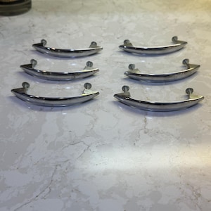May include: Six shiny, curved metal cabinet pulls with a polished finish. Each pull has a rounded handle and two small, circular mounting points. The pulls are arranged on a light-colored surface, suggesting a hardware or home decor theme.