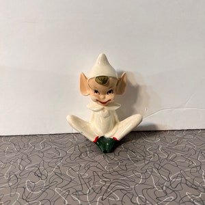 Può includere: Una statuetta di elfo in ceramica bianca con scarpe verdi e un cappello a punta. L'elfo è seduto a gambe incrociate e ha un'espressione maliziosa sul viso.