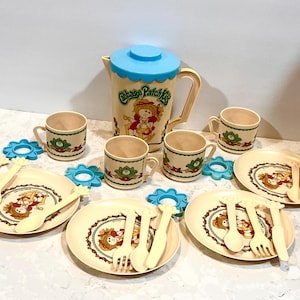 May include: A vintage Cabbage Patch Kids tea set with a blue pitcher, four cups, four saucers, and four plates. The pitcher has a blue lid and a Cabbage Patch Kids character on the front. The cups, saucers, and plates have a floral design and a Cabbage Patch Kids character on the front.