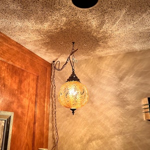 May include: A gold-colored, round, hanging light fixture with a chain and a decorative pattern. The light is on and illuminating the room.